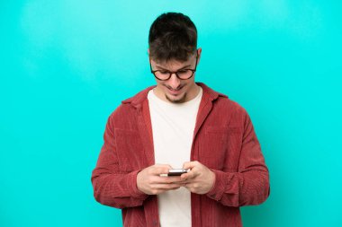 Mavi arka planda izole genç beyaz adam cep telefonuyla bir mesaj gönderiyor.
