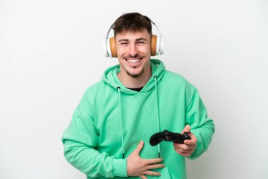 Beyaz arka planda izole edilmiş bir bilgisayar oyunu kontrolörüyle oynayan yakışıklı genç adam çok gülümsüyor.