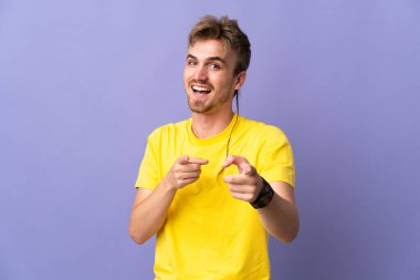 Mor arka planda soyutlanmış yakışıklı sarışın adam şaşırmış ve ön tarafı işaret ediyor.