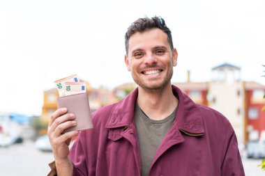 Açık havada genç beyaz bir adam elinde cüzdanla mutlu bir ifadeyle.