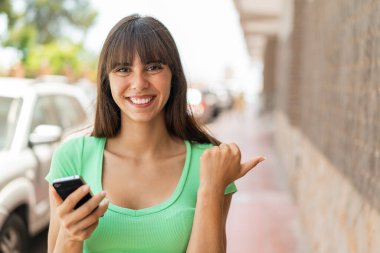 Açık havada cep telefonu kullanan ve yan tarafı işaret eden genç bir kadın.