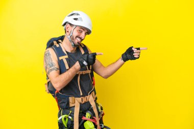 Genç beyaz dağcı sarı zemin üzerinde izole edilmiş bir şekilde parmak göstererek bir ürün sunuyor.