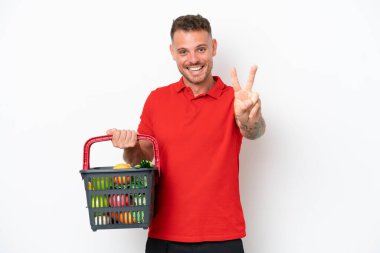 Beyaz arka planda izole edilmiş yiyecek dolu bir sepet taşıyan beyaz bir adam gülümsüyor ve zafer işareti gösteriyor.