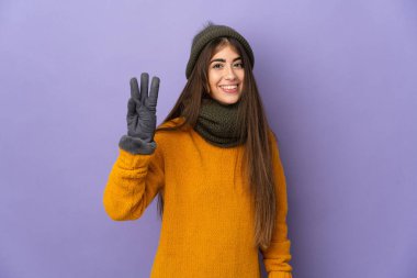 Mor arka planda izole edilmiş beyaz şapkalı genç kız mutlu ve parmaklarıyla üçe kadar sayıyor.