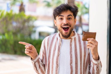 Açık havada cüzdan tutan yakışıklı bir Arap şaşırmış ve parmağıyla onu işaret ediyor.