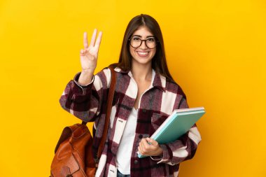 Sarı arka planda izole edilmiş genç bayan mutlu ve parmaklarıyla üçe kadar sayıyor.