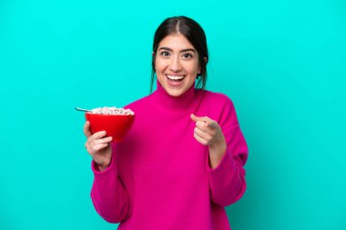 Mavi arka planda bir kase mısır gevreği tutan beyaz bir kadın şaşırmış ve ön tarafı işaret ediyor.