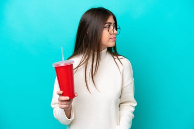 Mavi arka planda izole edilmiş gazoz içen genç beyaz kadın yan tarafa bakıyor.