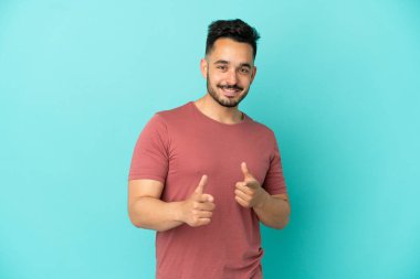 Mavi arka planda izole edilmiş genç beyaz adam ön tarafı işaret ediyor ve gülümsüyor.