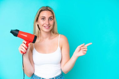 Mavi arka planda izole edilmiş saç kurutma makinesini tutan beyaz bir kadın şaşırmış ve parmağını kenarı işaret ediyor.