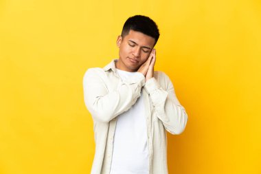 Genç Ekvadorlu, sarı arka planda izole edilmiş bir şekilde uyku jesti yapıyor.