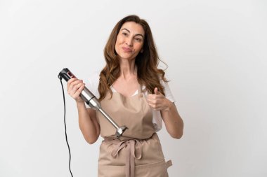 Beyaz arka planda izole edilmiş el blenderi kullanan orta yaşlı beyaz bir kadın baş parmak işareti yapıyor.