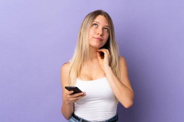 Mor arka planda izole edilmiş cep telefonu kullanan genç sarışın bir kadın bir fikir düşünüyor.