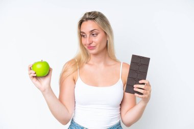 Beyaz arka planda izole olmuş genç beyaz bir kadın bir elinde çikolata tableti, diğer elinde elma tutarken kuşkuları var.