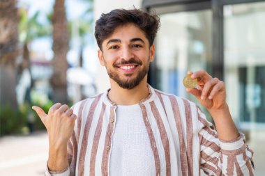 Dışarıda Bitcoin tutan yakışıklı bir Arap, bir ürünü sunmak için yan tarafı işaret ediyor.