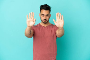 Mavi arka planda izole edilmiş genç beyaz adam dur hareketi yapıyor ve hayal kırıklığına uğruyor.