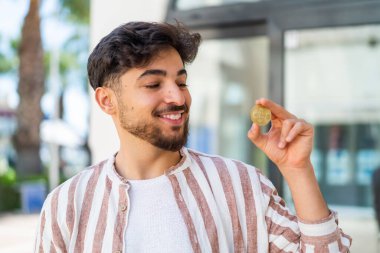 Yakışıklı bir Arap açık havada Bitcoin tutuyor, neşeli bir ifadeyle.