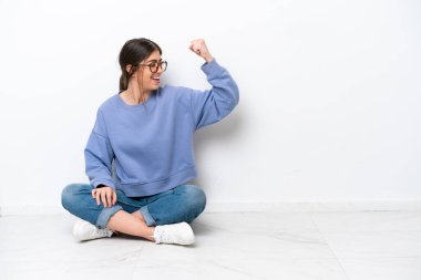 Beyaz arka planda izole bir şekilde yerde oturan beyaz bir kadın güçlü bir jest yapıyor.