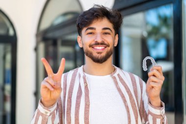 Yakışıklı bir Arap, dışarıda görünmez diş telleri tutuyor. Gülümsüyor ve zafer işareti gösteriyor.