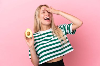 Avokado tutan beyaz bir kadın pembe arka planda izole edilmiş, gülümsüyor.