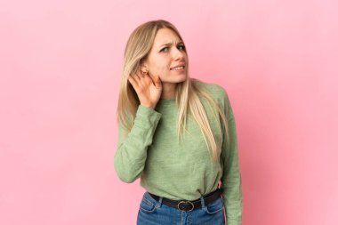 Pembe arka planda izole edilmiş genç sarışın bir kadın. Kulağına elini sürerek bir şeyler dinliyor.