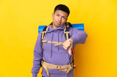 Sarı arka planda izole edilmiş büyük sırt çantalı genç dağcı, olumsuz ifadeli baş parmağını gösteriyor.