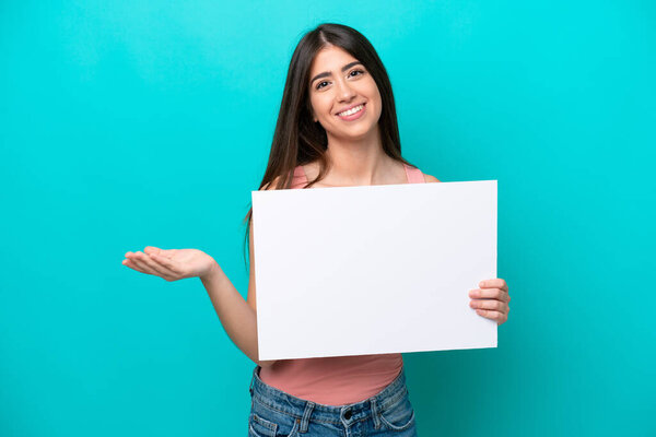 Молодая белая женщина, изолированная на синем фоне, держит пустой плакат с удивленным выражением