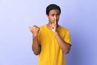 Siyahi örgülü genç Afrikalı Amerikalı adam, mor arka planda izole edilmiş, kenarı işaret ediyor ve sessizlik hareketi yapıyor.