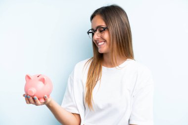 Elinde kumbara tutan beyaz bir kadın mavi arka planda mutlu bir ifadeyle izole edilmiş.