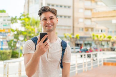Genç yakışıklı adam dışarıda cep telefonu kullanıyor, mutlu bir ifadeyle.