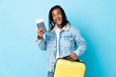 Saçları mavi arka planda izole edilmiş, bavul ve pasaportlu genç Afrikalı Amerikalı bir adam.