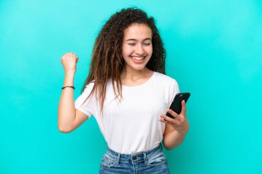 Genç Arap kadın mavi arka planda cep telefonu kullanarak ve zafer jesti yaparak izole edildi.