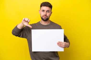 Sarı arka planda izole edilmiş genç beyaz bir adam boş bir levha tutuyor ve kötü sinyal veriyor.