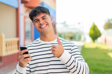 Genç İspanyol adam dışarıda cep telefonu kullanıyor. Başparmakları havada çünkü iyi bir şey oldu.