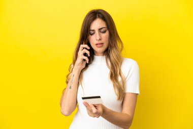 Beyaz kadın sarı arka planda izole edilmiş cep telefonuyla kredi kartıyla alışveriş yapıyor.
