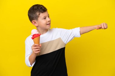 Kornet dondurması olan küçük beyaz bir çocuk sarı arka planda izole edilmiş bir şekilde baş parmak hareketi yapıyor.