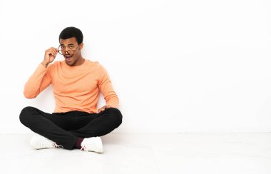 Afrika kökenli Amerikalı bir adam, gözlüklü ve şaşırmış bir şekilde tecrit edilmiş bir arka planda oturuyor.