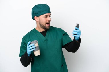 Yeşil üniformalı bir cerrah beyaz arka planda izole edilmiş. Elinde kahve ve cep telefonu var.