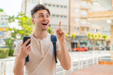 Dışarıda cep telefonu kullanan genç yakışıklı bir adam çözümü bulmak için parmağını kaldırıyor.