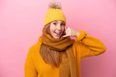 Kışlık ceket giyen genç kızıl saçlı kadın pembe arka planda izole edilmiş telefon jesti yapıyor. Bana geri dönüş işareti ver.