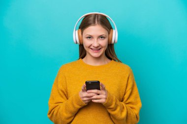 Genç Rus kadın mavi arka planda izole edilmiş. Cep telefonuyla müzik dinliyor ve ön tarafa bakıyor.
