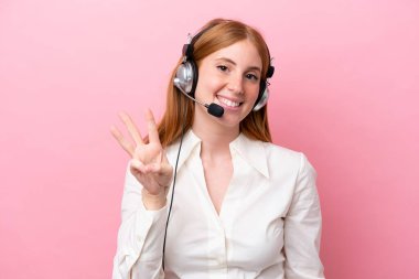 Pembe arka planda izole edilmiş kulaklıkla çalışan pazarlamacı kızıl saçlı kadın mutlu ve parmaklarıyla üçe kadar sayıyor.