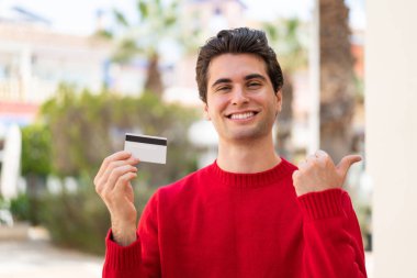 Dışarıda elinde bir kredi kartı olan genç yakışıklı bir adam bir ürünü sunmak için kenarı işaret ediyor.