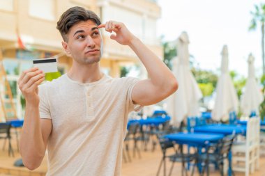 Açık havada kredi kartı tutan genç yakışıklı adamın şüpheleri var ve kafası karışmış bir yüz ifadesi var.