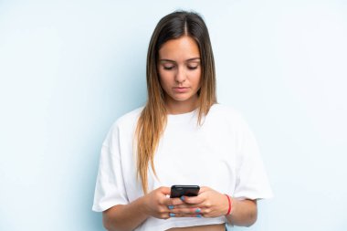 Genç beyaz kadın mavi arka planda cep telefonu kullanıyor.