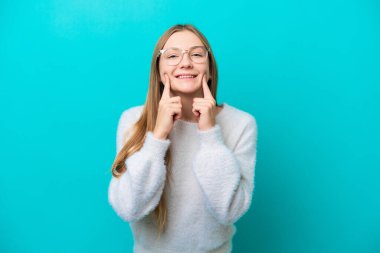 Mavi arka planda izole edilmiş genç Rus kadın mutlu ve hoş bir ifadeyle gülümsüyor.