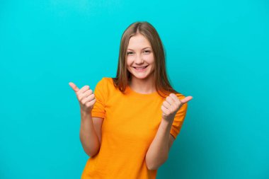 Mavi arka planda izole edilmiş genç bir Rus kadın. Baş parmakları havada ve gülümsüyor.