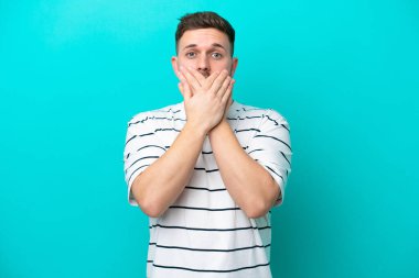 Mavi arka planda izole edilmiş genç beyaz adam elleriyle ağzını kapatıyor.