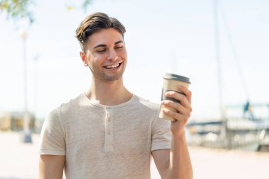Genç yakışıklı bir adam açık havada elinde kahveyle neşeli bir ifade ile geziyor.