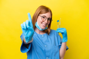Genç kızıl saçlı dişçi kadın sarı arka planda izole edilmiş ve parmağını kaldırıyor.